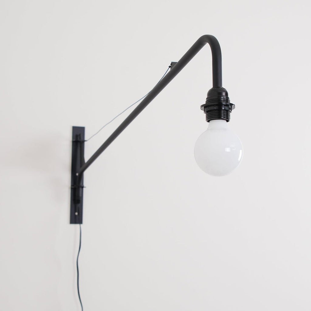 Close view of the black back plate, arm and round white globe bulb of this Clérachic wall light, with a black cord below.