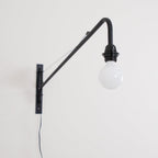 Close view of the black back plate, arm and round white globe bulb of this Clérachic wall light, with a black cord below.
