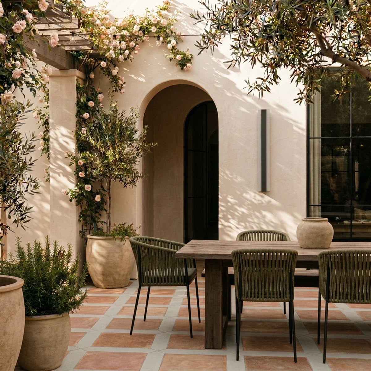 By day, the black column sconce lights a stucco wall beside a rose-covered pergola over an outdoor dining table, Clérachic lifestyle shot.