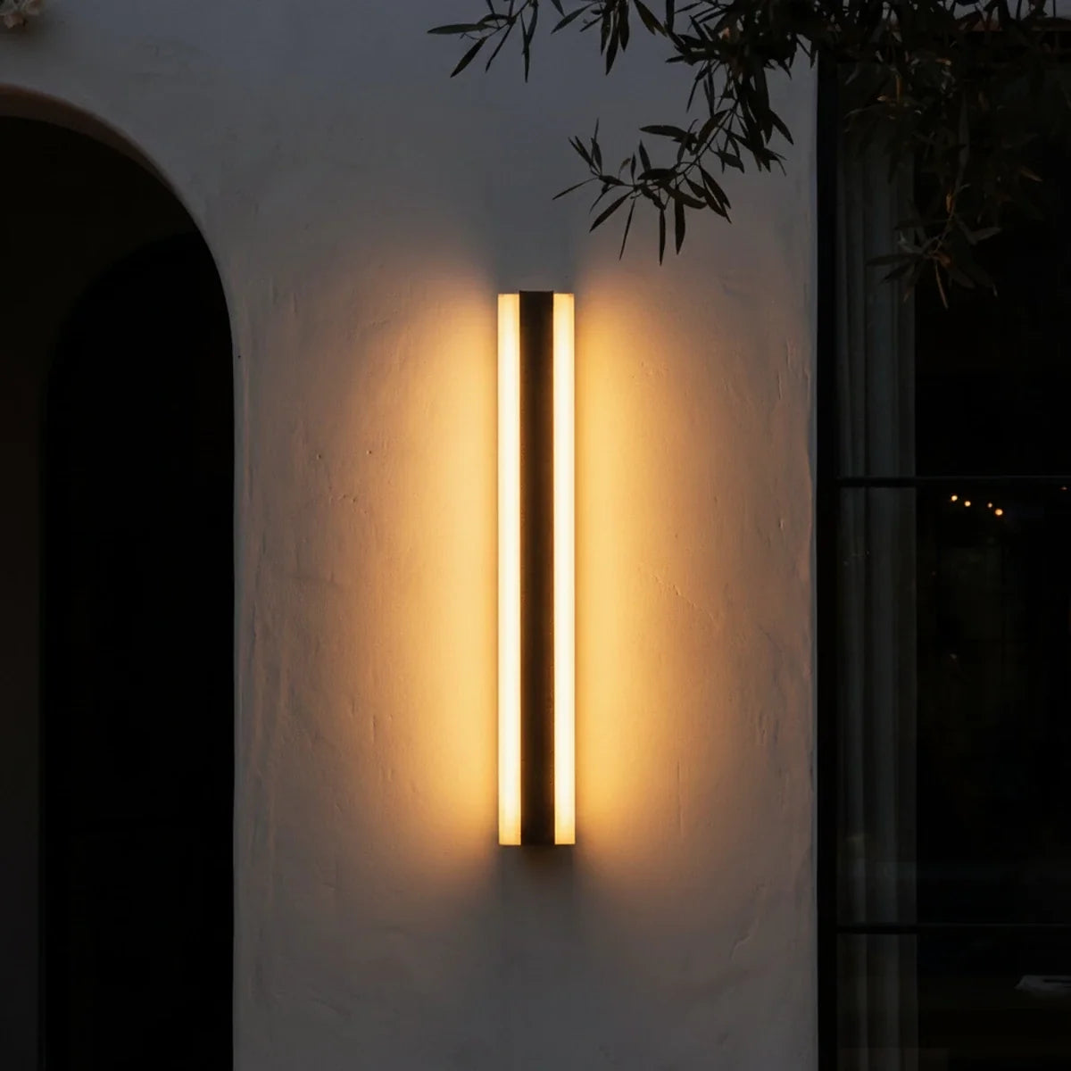 At night, a single black column sconce glows beside an archway under olive branches in this Clérachic lifestyle shot.