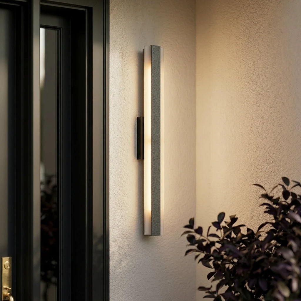 A close-up shows a raw aluminium-finish column sconce mounted beside a dark green door at dusk, Clérachic detail shot.