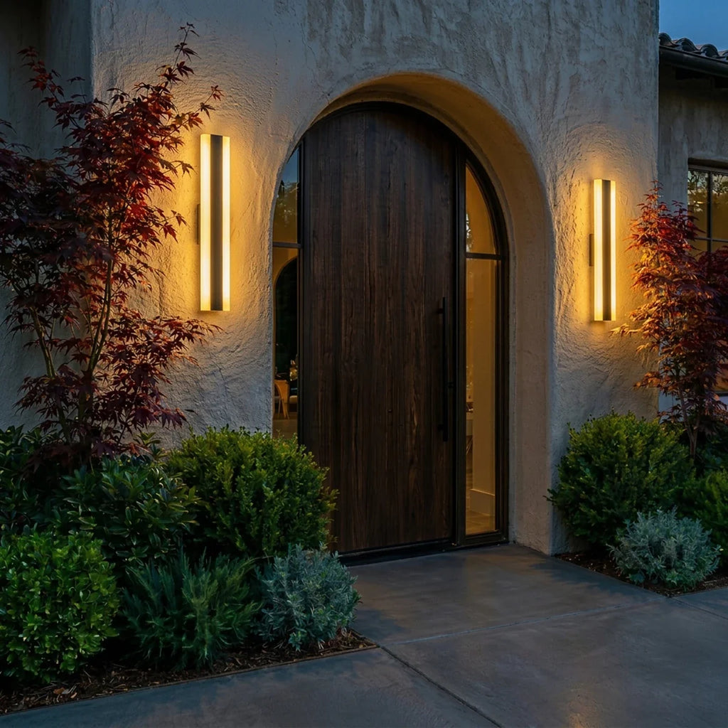 At night, the same two black column sconces glow warm beside the arched wood door and maple trees, Clérachic lifestyle shot.