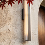 A close-up on a raw aluminium-finish column sconce glowing beside red maple leaves near an arched doorway, Clérachic detail shot.