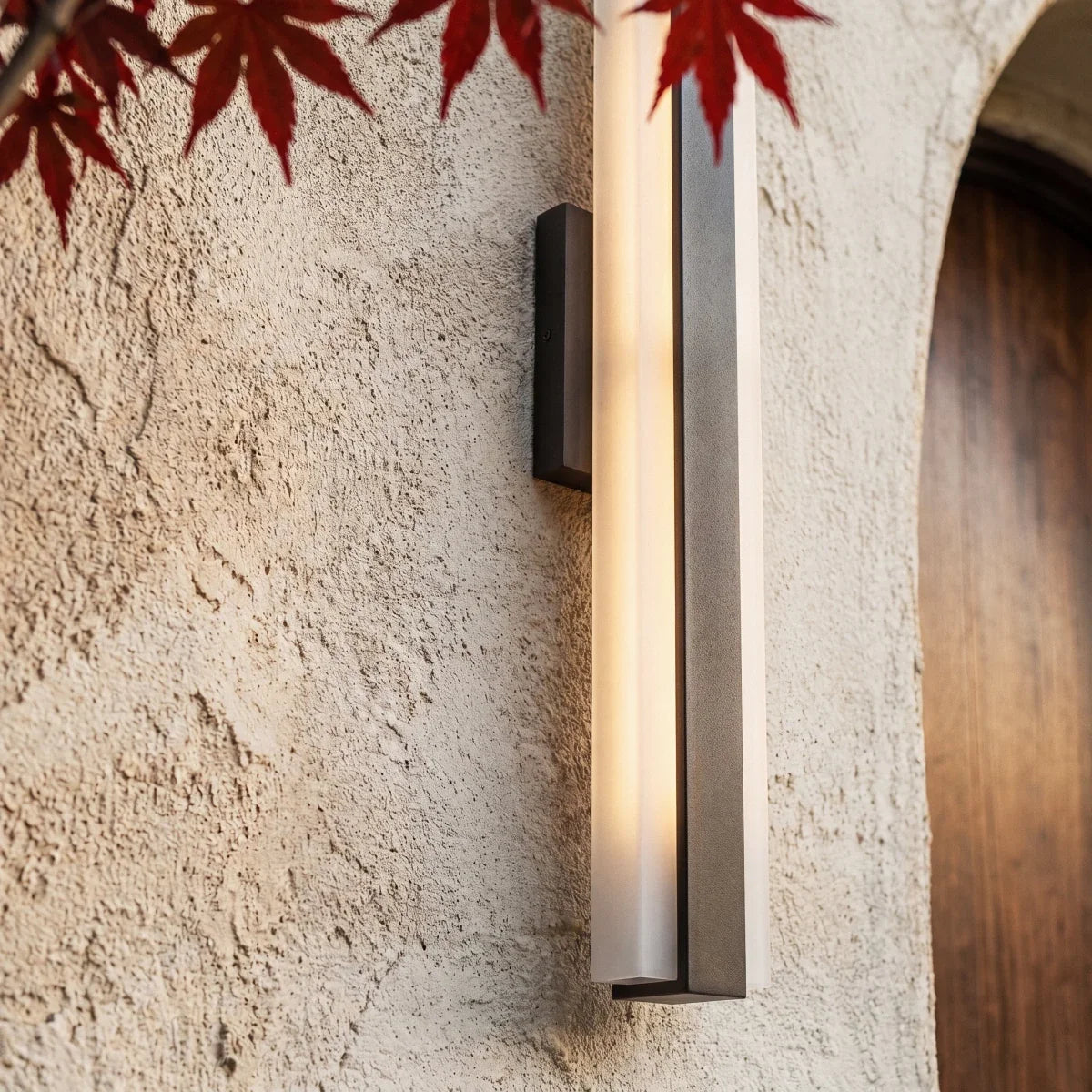 A close-up on a raw aluminium-finish column sconce glowing beside red maple leaves near an arched doorway, Clérachic detail shot.