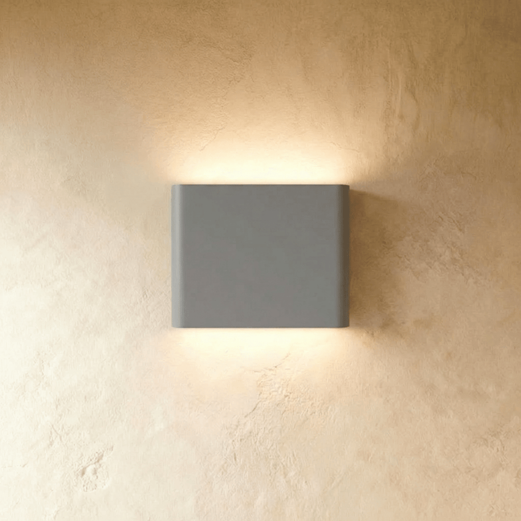 A compact grey up-down wall sconce washes warm light both ways on a plaster wall in this Clérachic hero shot.
