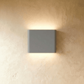 A compact grey up-down wall sconce washes warm light both ways on a plaster wall in this Clérachic hero shot.