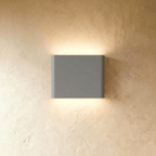 A compact grey up-down wall sconce washes warm light both ways on a plaster wall in this Clérachic hero shot.