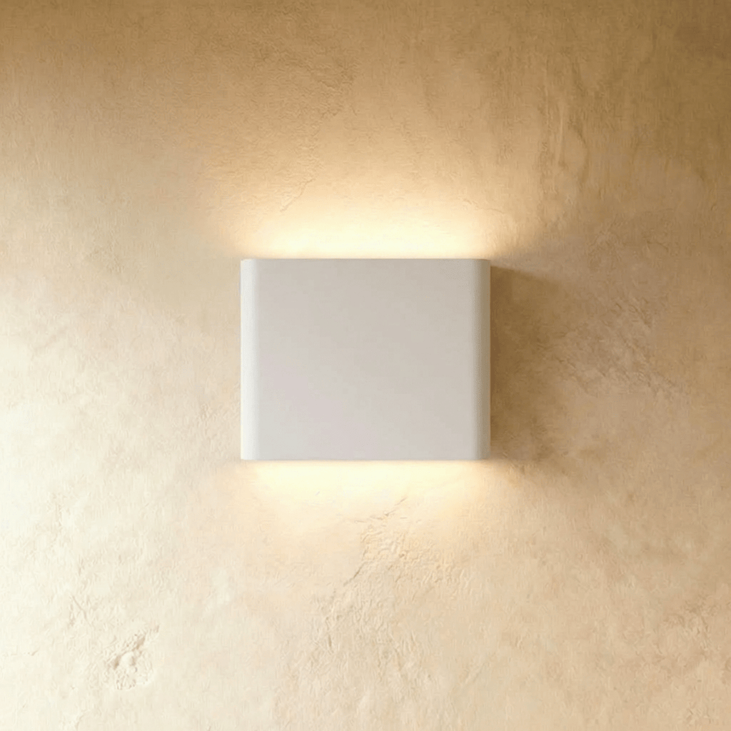 A white up-down wall sconce glows warm above and below its rounded body on a plaster wall, Clérachic hero shot.