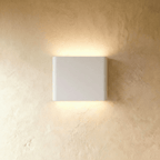 A white up-down wall sconce glows warm above and below its rounded body on a plaster wall, Clérachic hero shot.