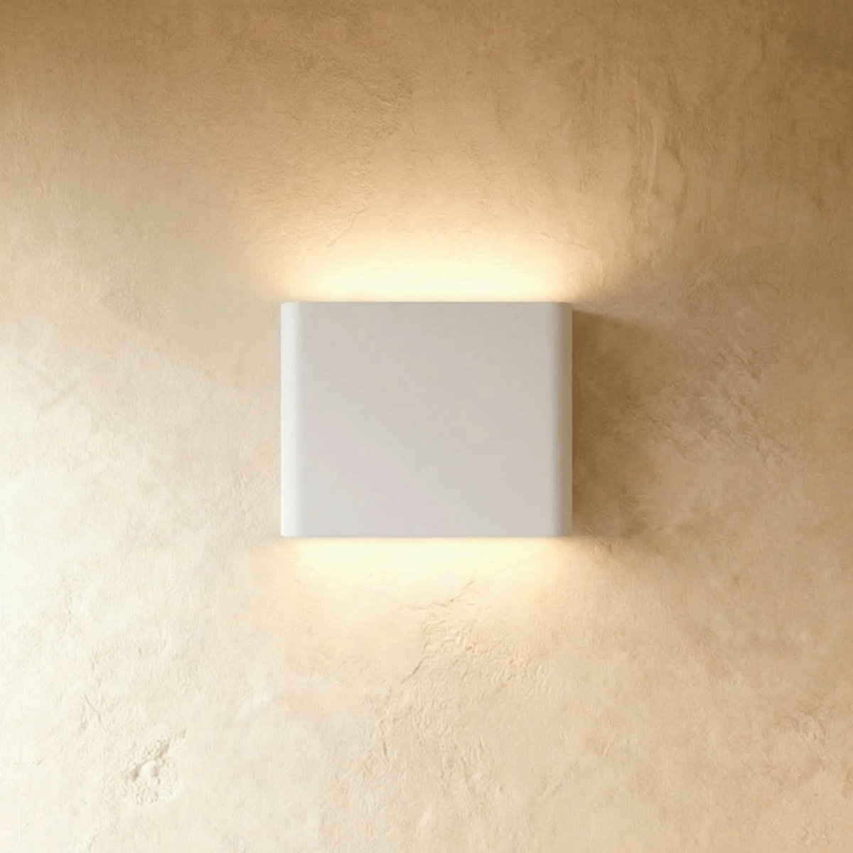 A white up-down wall sconce glows warm above and below its rounded body on a plaster wall, Clérachic hero shot.