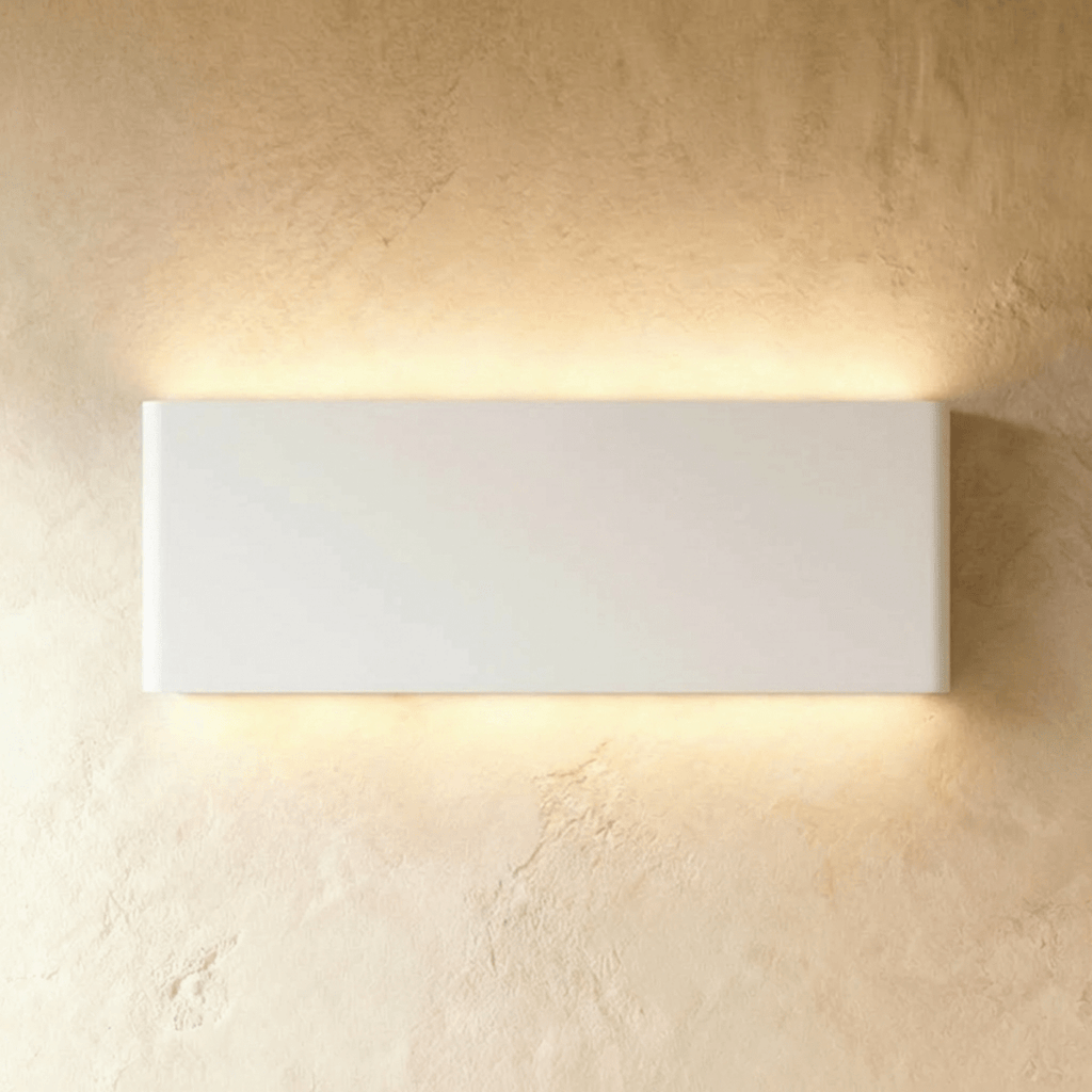 The largest white up-down sconce glows warm along a long rectangular body against plaster, Clérachic hero shot.