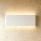 The largest white up-down sconce glows warm along a long rectangular body against plaster, Clérachic hero shot.