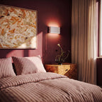 A white up-down sconce glows above a burl-wood nightstand in a deep-rose bedroom with striped bedding, Clérachic lifestyle shot.