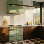 A Clérachic black walnut pendant bar glows warmly above a sage green range hood in this kitchen scene.