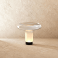 Clérachic mushroom-shaped glass table lamp lit warm inside its frosted white stem, with a clear dish top and black base.