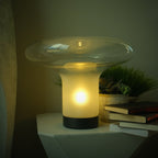 Clérachic mushroom glass table lamp glowing warm on a white side table beside books and a potted succulent.