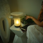 Clérachic mushroom glass table lamp lit on a white faceted side table, with a hand reaching toward it and a succulent beside.