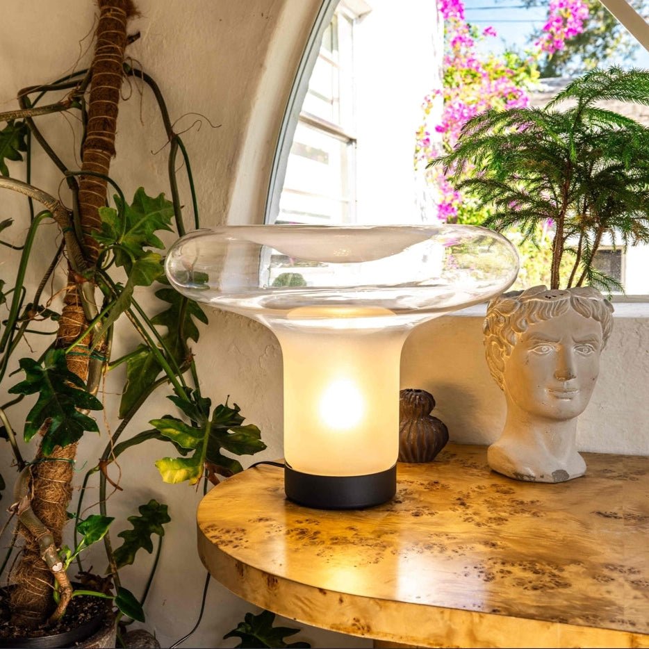 Clérachic mushroom glass table lamp glowing on a burl wood table by an arched window, with plants and a bust-shaped planter.