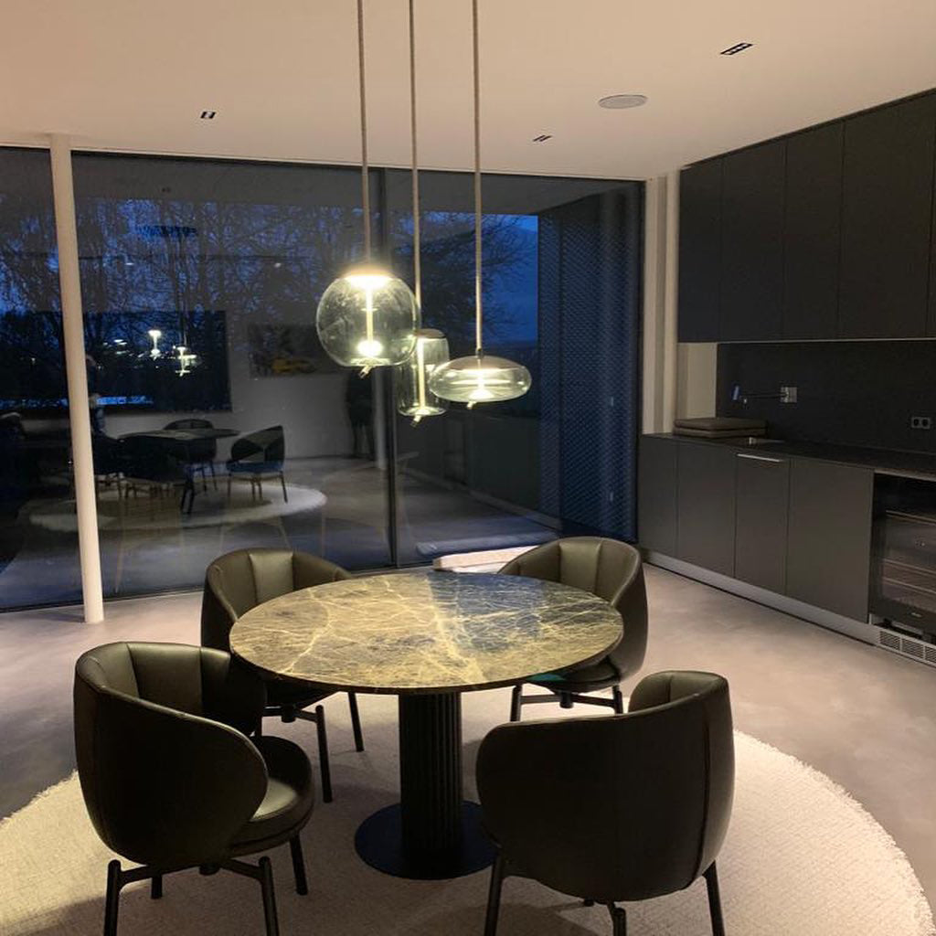 Lit at dusk, a round glass Clérachic pendant and two smaller ones hang above a round marble-topped table with dark leather armchairs.