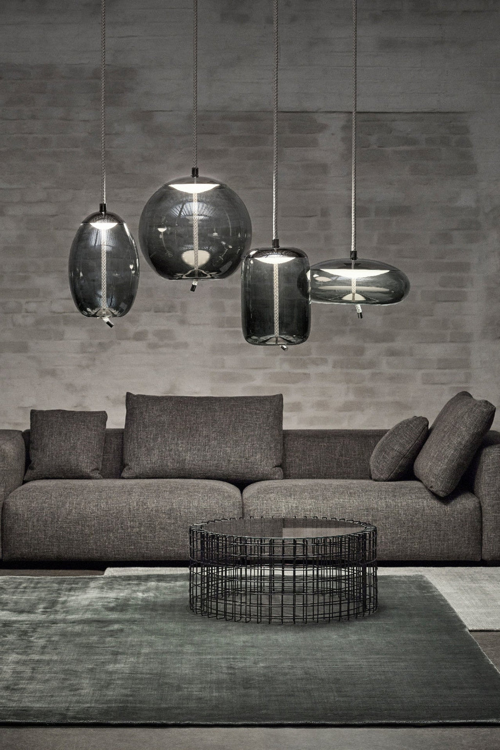 Four smoke-grey glass Clérachic pendants in round, tall and flat shapes hang at different heights against a grey brick wall above a sofa.