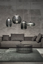 Four smoke-grey glass Clérachic pendants in round, tall and flat shapes hang at different heights against a grey brick wall above a sofa.