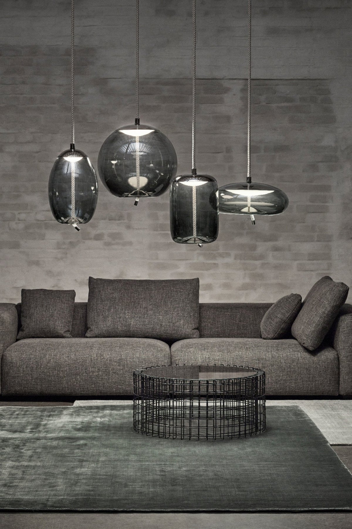 Four smoke-grey glass Clérachic pendants in round, tall and flat shapes hang at different heights against a grey brick wall above a sofa.