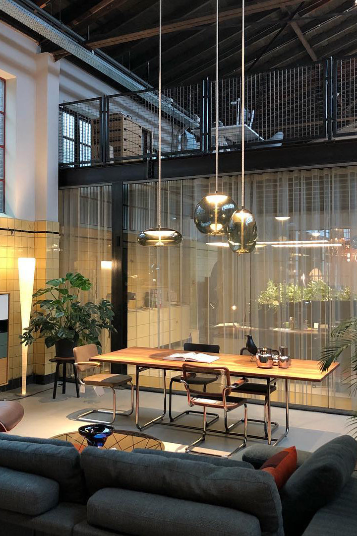 In a tall industrial loft, three small glass Clérachic pendants glow warm above a wooden dining table and a dark sofa.