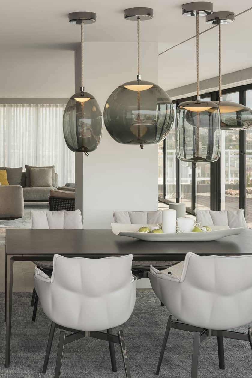 Round and tall smoke-grey Clérachic glass pendants hang from dark ceiling canopies over a dining table with white chairs.