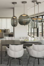 Round and tall smoke-grey Clérachic glass pendants hang from dark ceiling canopies over a dining table with white chairs.