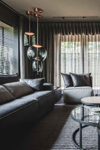 Smoke-grey glass Clérachic pendants hang from copper-toned canopies in a dark living room with heavy curtains and a leather sofa.