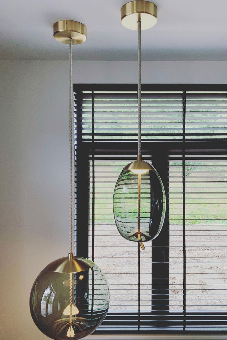 Two glass Clérachic pendants hang at different heights from brass-toned canopies in front of a black-framed window.