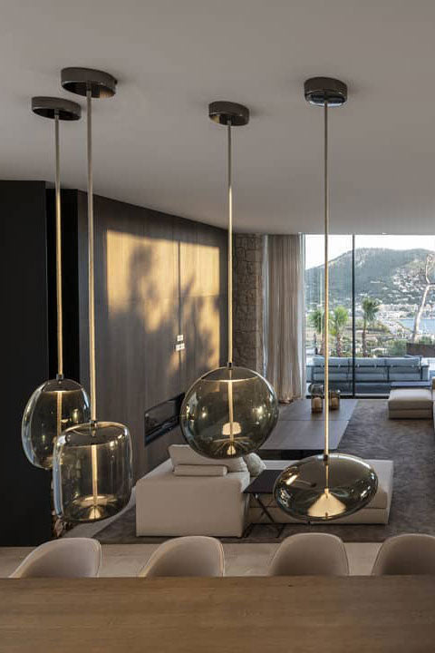 Four lit glass Clérachic pendants with brass-toned fittings hang above a low living area with a sea view beyond the glass wall.