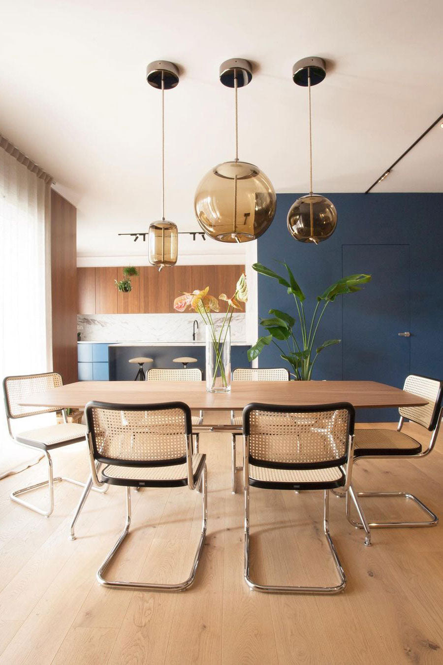 Amber glass Clérachic pendants in round, tall and oval shapes hang over a wooden dining table with cane-seated chairs and a blue wall behind.