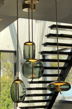 Looking up from a staircase, a cluster of smoke-grey glass Clérachic pendants in oval, round and flat shapes glow warm.