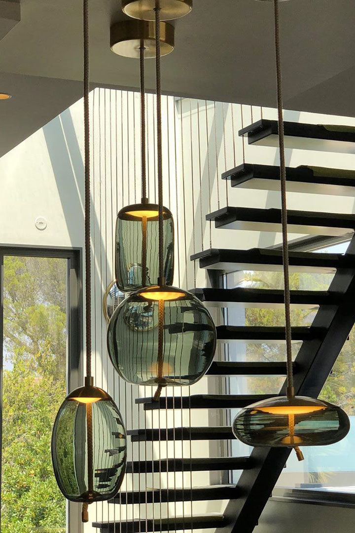 Looking up from a staircase, a cluster of smoke-grey glass Clérachic pendants in oval, round and flat shapes glow warm.