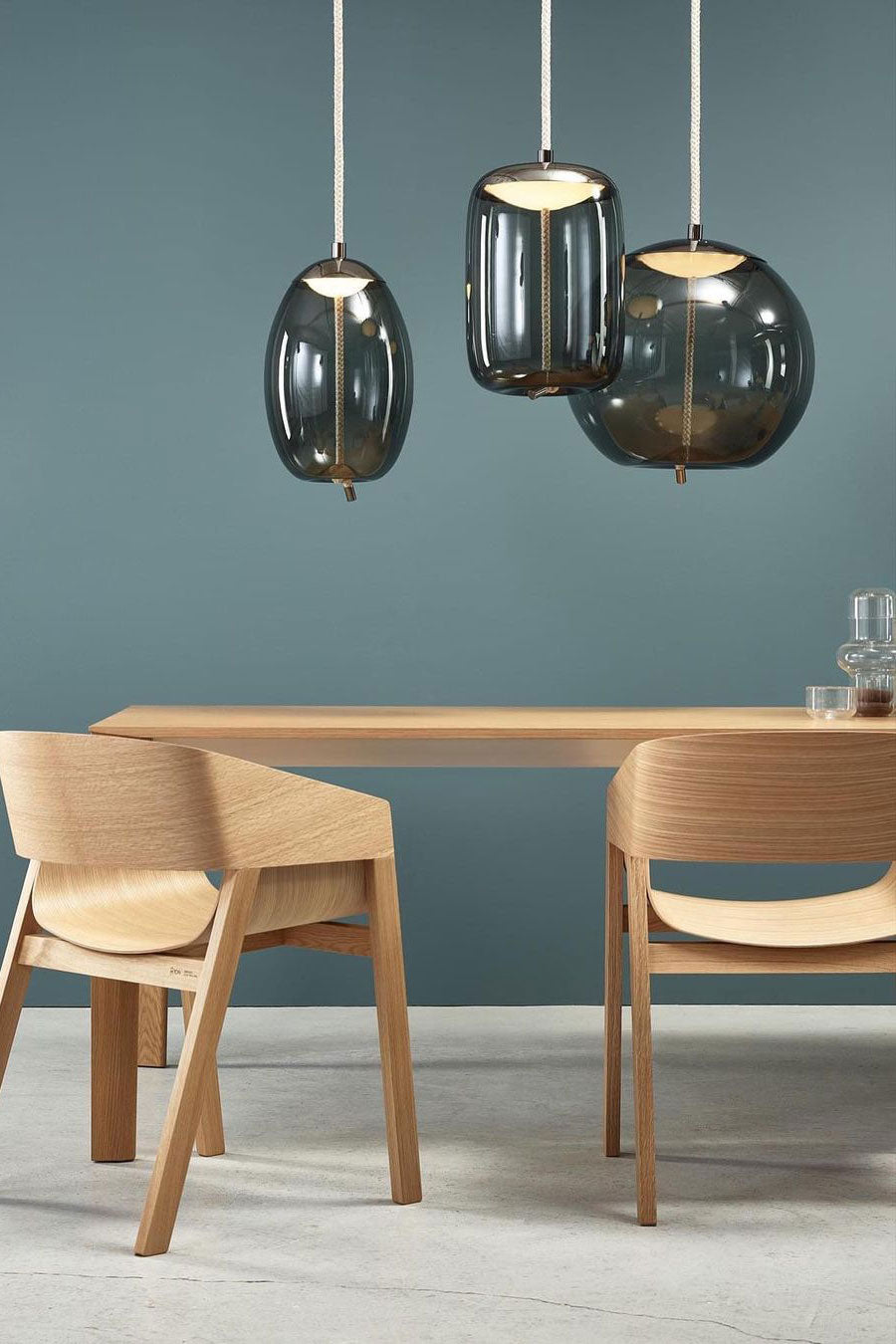 Three smoke-grey glass Clérachic pendants hang against a blue-grey wall above a wooden table with two pale wooden chairs.