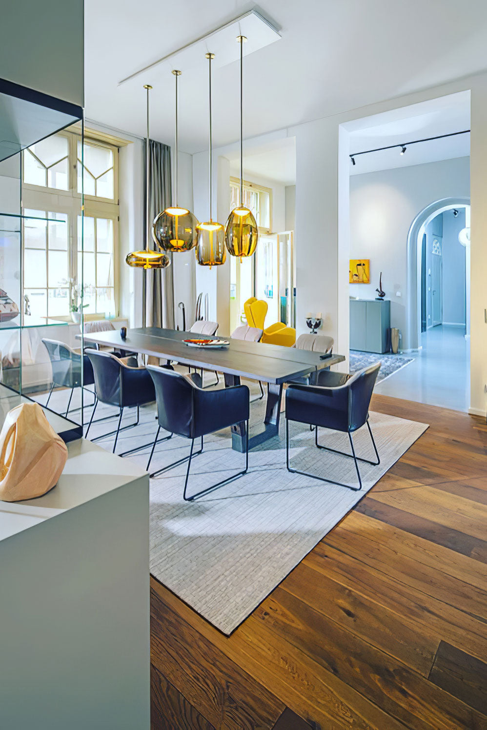 A row of amber glass Clérachic pendants hangs from a long ceiling plate above a grey dining table with blue chairs.