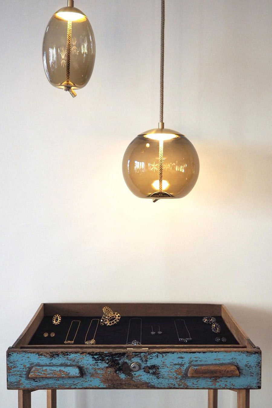 A round amber-smoke glass Clérachic pendant glows warm beside a smaller oval one, above a worn blue-painted wooden table.