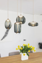 Four glass Clérachic pendants in grey-green tints hang in a white room above a wooden table with a vase of yellow tulips.