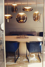 Amber-tinted glass Clérachic pendants in round, flat and oval shapes hang above an oval dining table with deep blue chairs.