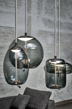 Close view of three smoke-grey glass Clérachic pendants with braided cords, hanging against a grey brick wall.