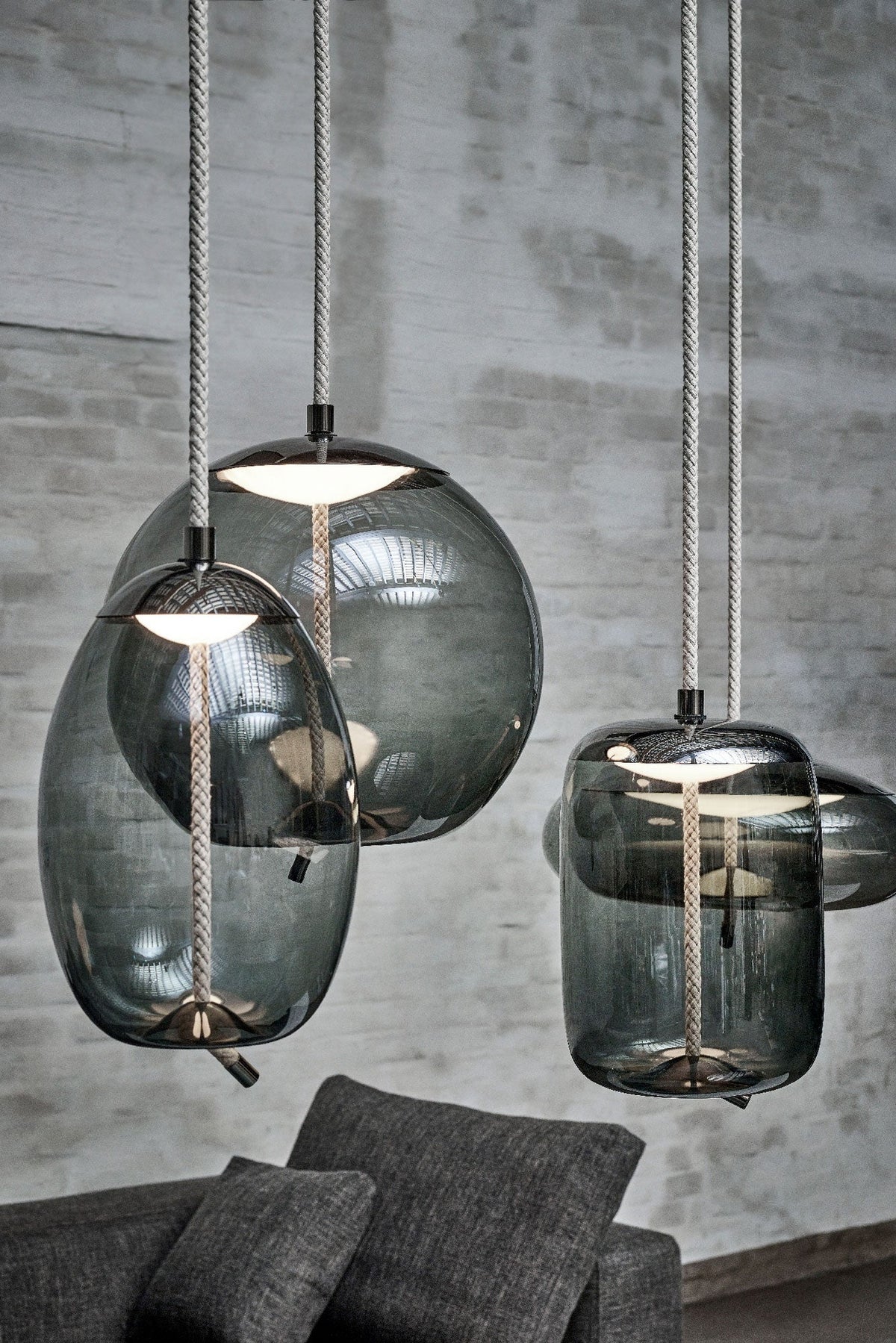 Close view of three smoke-grey glass Clérachic pendants with braided cords, hanging against a grey brick wall.