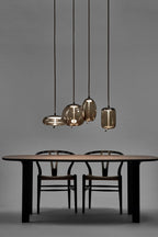 Four small Clérachic glass pendants in graduated sizes hang low over a dark wooden table with two wishbone-style chairs on a grey backdrop.
