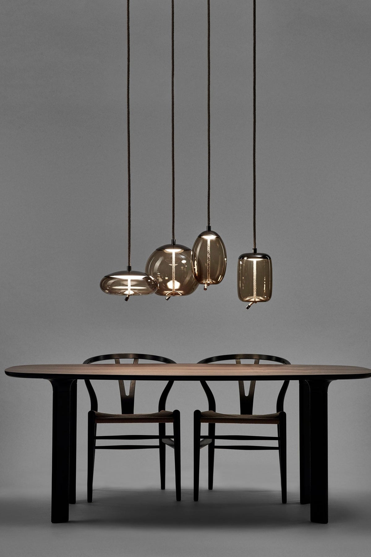 Four small Clérachic glass pendants in graduated sizes hang low over a dark wooden table with two wishbone-style chairs on a grey backdrop.