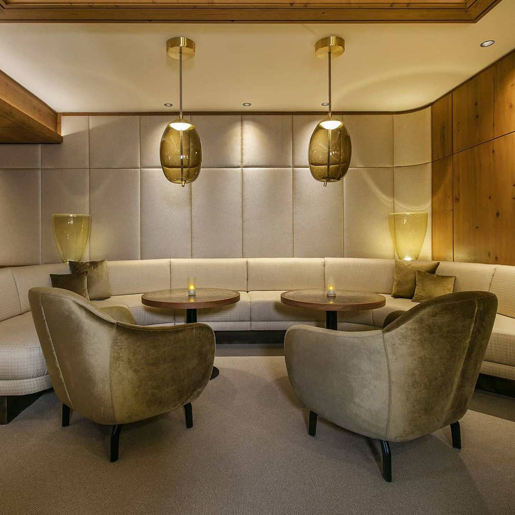 Two amber glass Clérachic pendants hang from brass-toned canopies in a hotel-style lounge with olive velvet armchairs and padded walls.