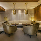 Two amber glass Clérachic pendants hang from brass-toned canopies in a hotel-style lounge with olive velvet armchairs and padded walls.