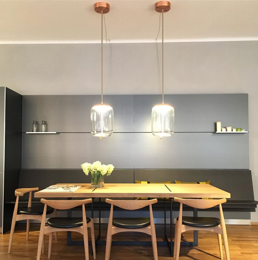 Two clear glass Clérachic pendants with copper-toned canopies hang above a pale wooden table with yellow flowers and four wooden chairs.