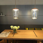 Two lit clear glass Clérachic pendants with copper-toned caps hang above a wooden table, against a dark grey panelled wall.