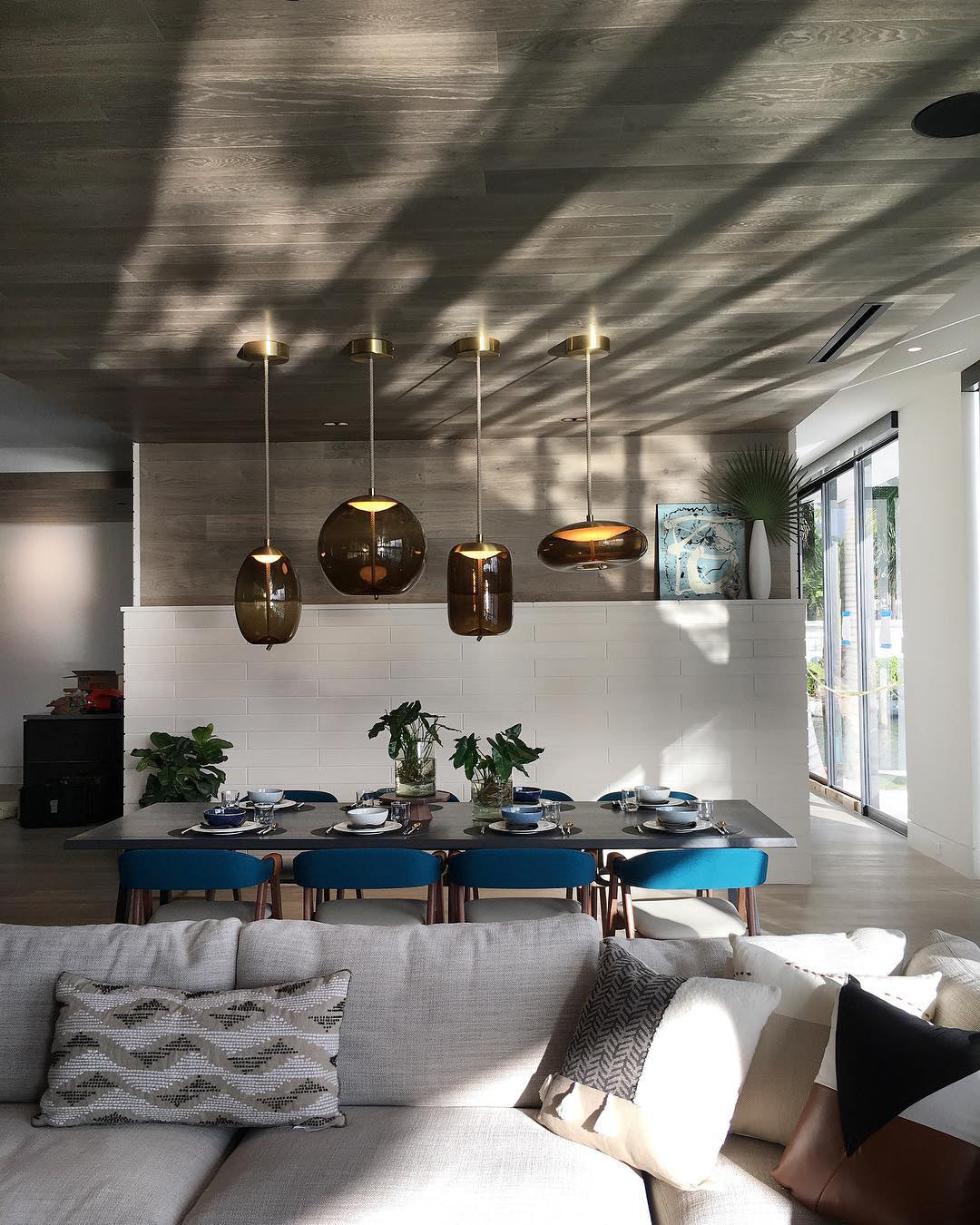 Four amber-brown glass Clérachic pendants hang in a bright open room above a set table, with a grey sofa in the foreground.