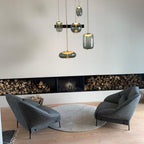 Smoke-grey glass Clérachic pendants hang from a black rail above two dark armchairs and a long fireplace with stacked logs.